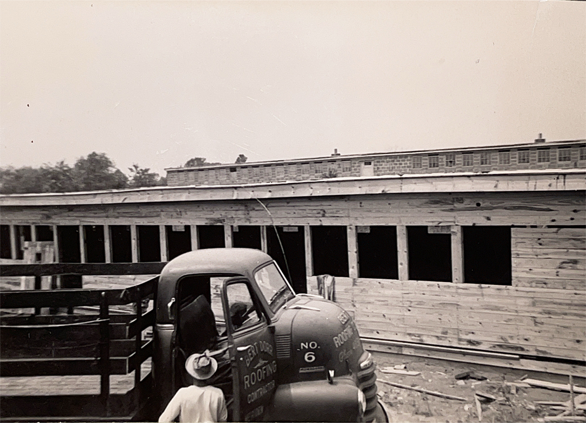 Black-and-white photo of a vintage work truck parked beside a long wooden building under construction