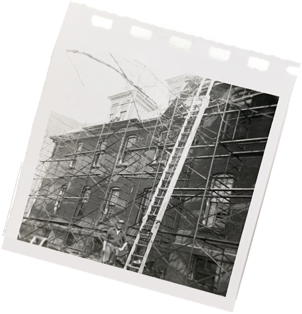 Black-and-white photo of a brick building surrounded by scaffolding with a ladder leaning against it