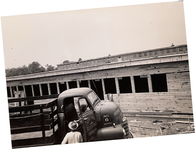 Black-and-white photo of a vintage work truck parked beside a long wooden building under construction